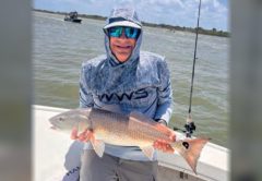 An angler in a hooded sun shirt and blue mirrored sunglasses holding a redfish on a boat, with open water behind him