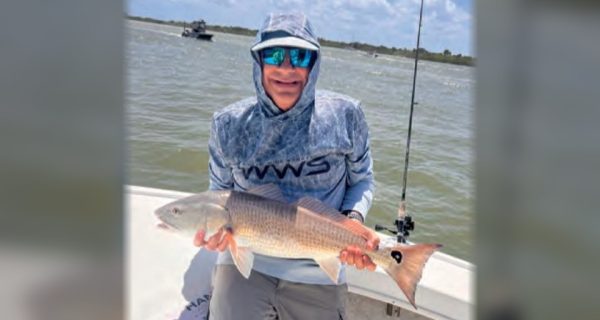 An angler in a hooded sun shirt and blue mirrored sunglasses holding a redfish on a boat, with open water behind him