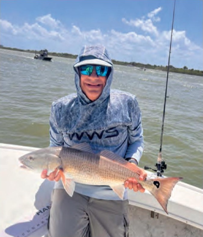 An angler in a hooded sun shirt and blue mirrored sunglasses holding a redfish on a boat, with open water behind him
