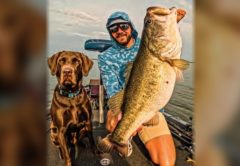 Capt. Tyler Woolcott in a hooded shirt and sunglasses holding up a giant largemouth bass on his boat, with his chocolate lab beside him