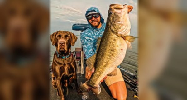 Capt. Tyler Woolcott in a hooded shirt and sunglasses holding up a giant largemouth bass on his boat, with his chocolate lab beside him