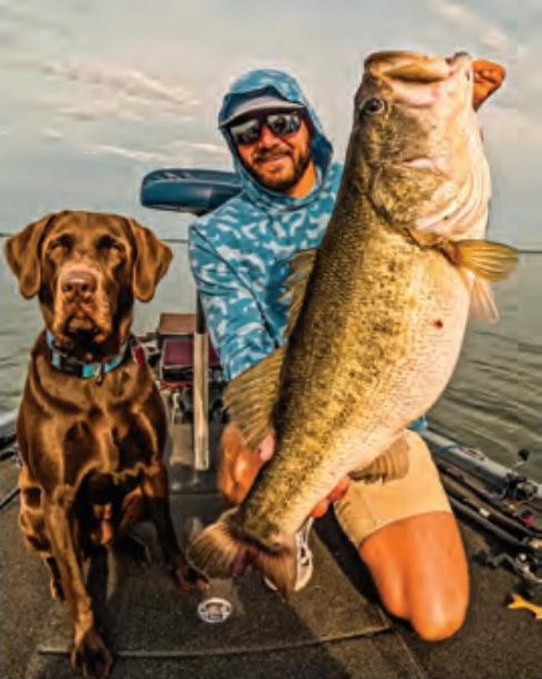 Capt. Tyler Woolcott in a hooded shirt and sunglasses holding up a giant largemouth bass on his boat, with his chocolate lab beside him