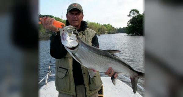 Capt. Barry Englehardt in a cap and vest holding up a tarpon on his boat, with a tree-lined river behind him