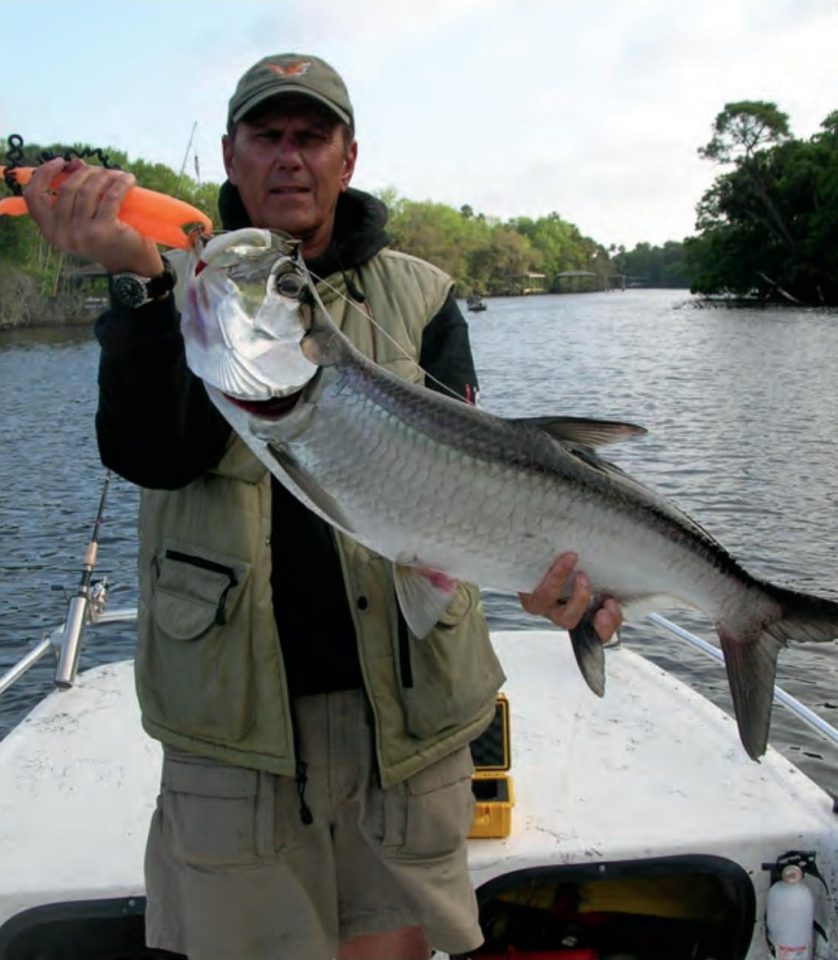 Capt. Barry Englehardt in a cap and vest holding up a tarpon on his boat, with a tree-lined river behind him