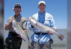 Two anglers holding up big speckled trout in front of open water under a clear blue sky