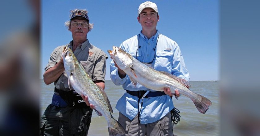 Two anglers holding up big speckled trout in front of open water under a clear blue sky