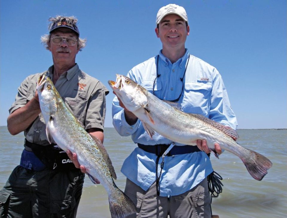 Two anglers holding up big speckled trout in front of open water under a clear blue sky