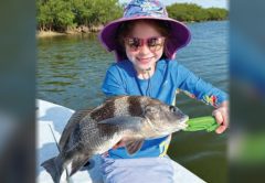 A smiling young angler in a floral sun hat and pink sunglasses holding a small black drum with a green fish gripper