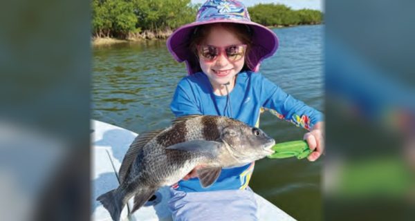 A smiling young angler in a floral sun hat and pink sunglasses holding a small black drum with a green fish gripper
