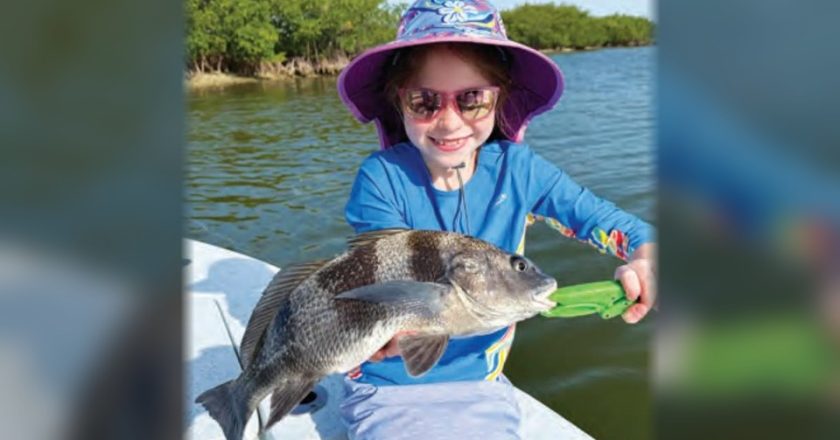 A smiling young angler in a floral sun hat and pink sunglasses holding a small black drum with a green fish gripper
