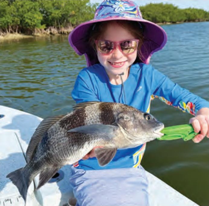 A smiling young angler in a floral sun hat and pink sunglasses holding a small black drum with a green fish gripper
