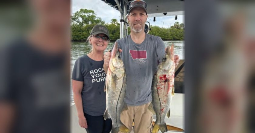A smiling couple on a boat in the river, the man holding up two snook, with mangroves behind them