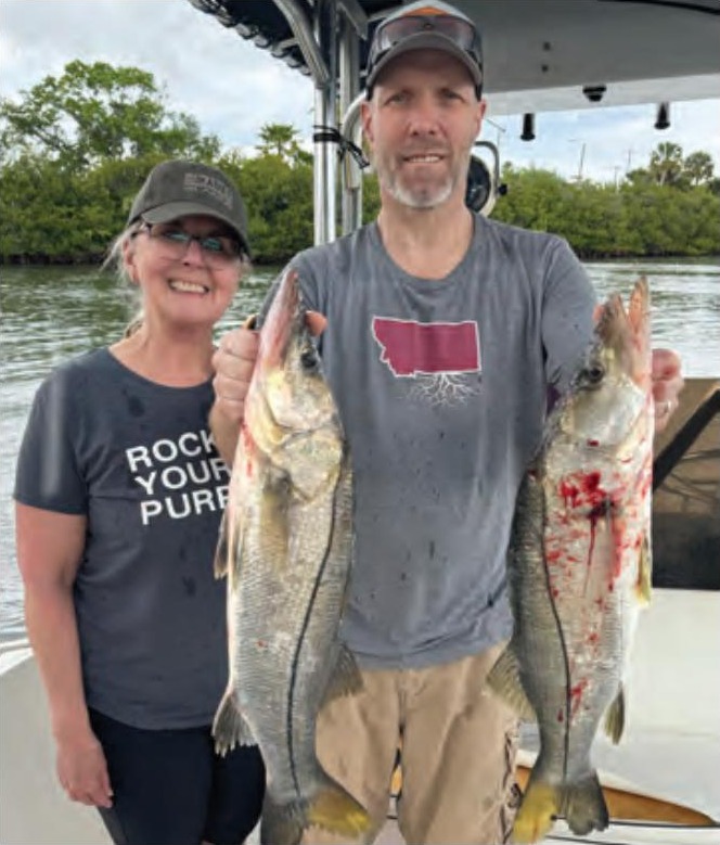 A smiling couple on a boat in the river, the man holding up two snook, with mangroves behind them