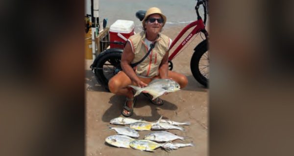 Marco Pompano crouching on the beach holding a pompano, with more pompano laid out on the sand and his red fat tire bike behind him