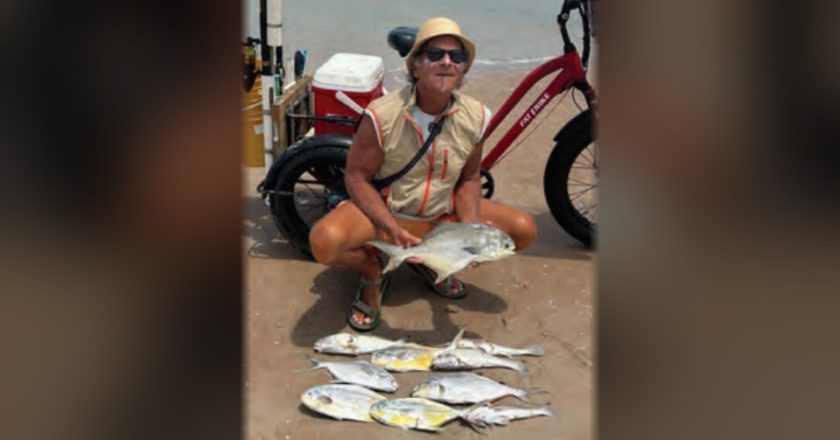 Marco Pompano crouching on the beach holding a pompano, with more pompano laid out on the sand and his red fat tire bike behind him