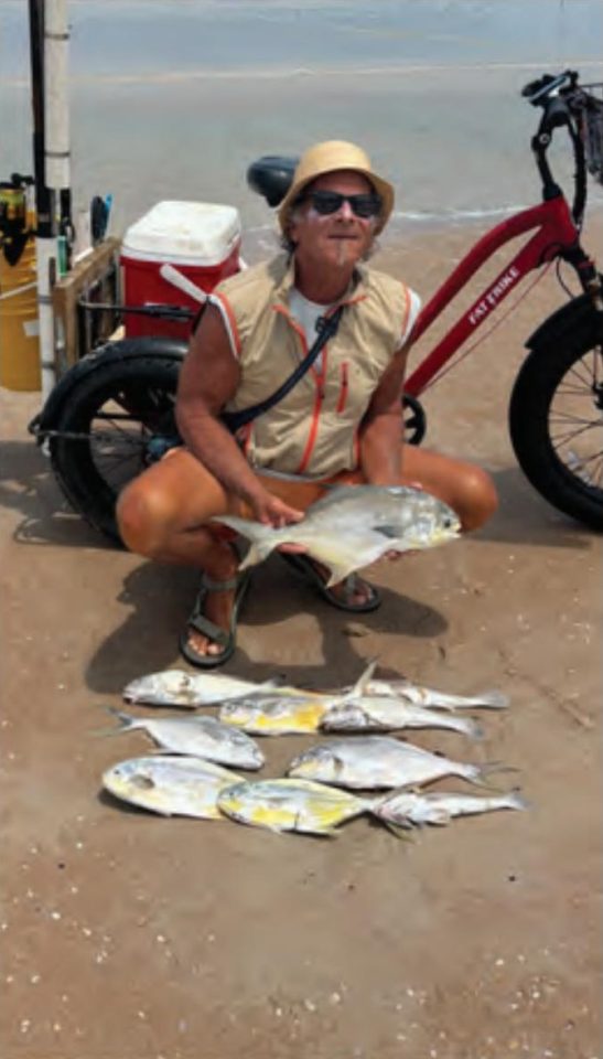 Marco Pompano crouching on the beach holding a pompano, with more pompano laid out on the sand and his red fat tire bike behind him