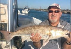 A smiling angler in a cap and sunglasses holding a redfish aboard a boat, with a rock jetty and the inlet behind him