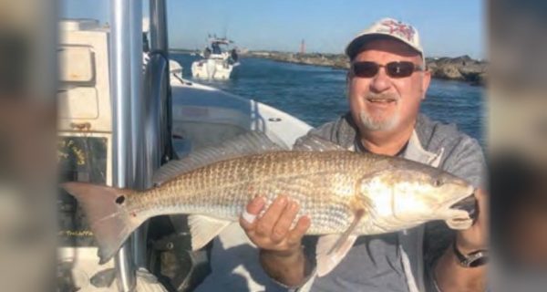 A smiling angler in a cap and sunglasses holding a redfish aboard a boat, with a rock jetty and the inlet behind him