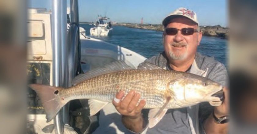 A smiling angler in a cap and sunglasses holding a redfish aboard a boat, with a rock jetty and the inlet behind him