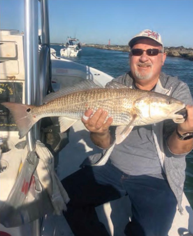 A smiling angler in a cap and sunglasses holding a redfish aboard a boat, with a rock jetty and the inlet behind him