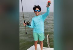 A smiling boy in blue sunglasses and a blue sun shirt holding up a popping cork on a boat, with a spotted seatrout hanging from his line
