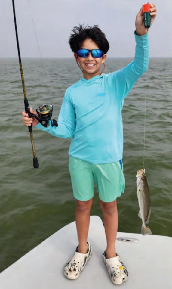 A smiling boy in blue sunglasses and a blue sun shirt holding up a popping cork on a boat, with a spotted seatrout hanging from his line