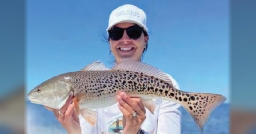 A smiling angler in a white cap and sunglasses holding a redfish covered in black spots, with the ocean behind her
