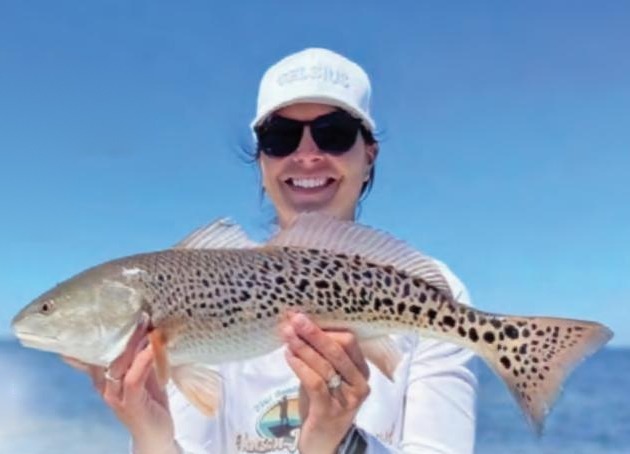 A smiling angler in a white cap and sunglasses holding a redfish covered in black spots, with the ocean behind her