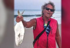 Marco Pompano in sunglasses and a red vest holding up a pompano by the tail on the beach
