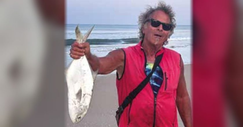 Marco Pompano in sunglasses and a red vest holding up a pompano by the tail on the beach