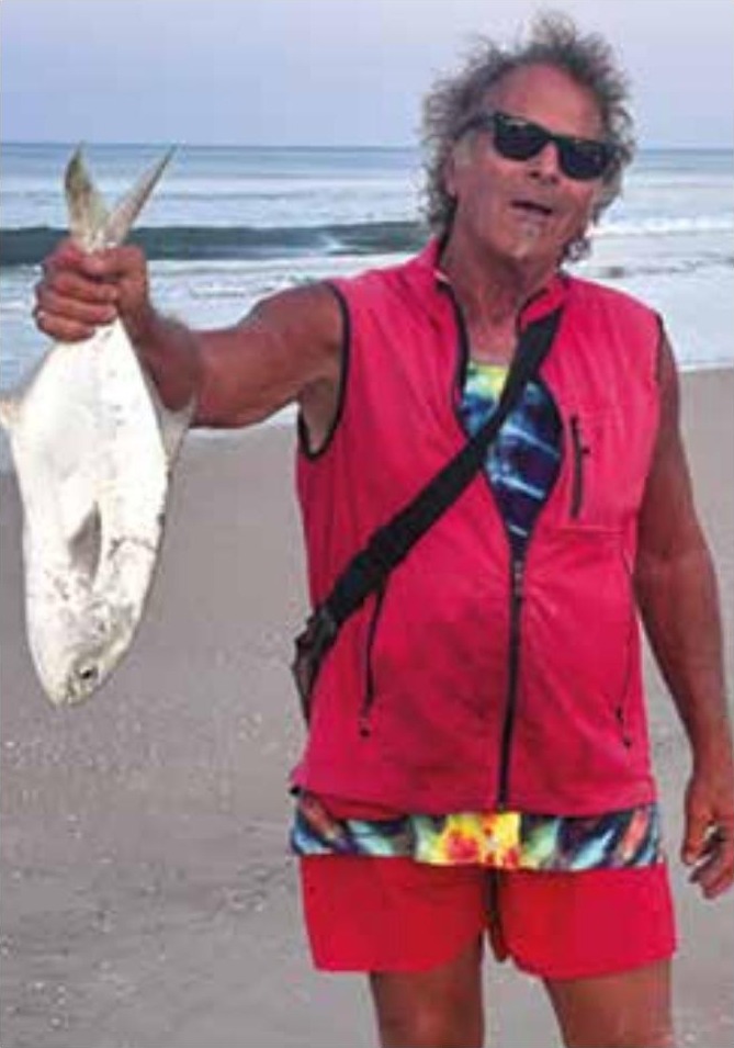 Marco Pompano in sunglasses and a red vest holding up a pompano by the tail on the beach