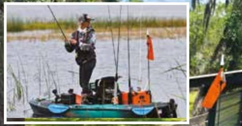 An angler standing and casting from a fishing kayak on a grassy lake, inset over a kayak on a stand loaded with a cooler, a safety flag, a paddle, and gear
