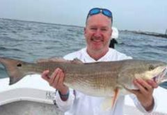 A smiling angler with sunglasses pushed up on his head, sitting on a cooler on a boat and holding a big redfish, with the inlet jetty behind him