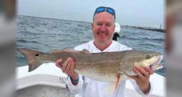 A smiling angler with sunglasses pushed up on his head, sitting on a cooler on a boat and holding a big redfish, with the inlet jetty behind him