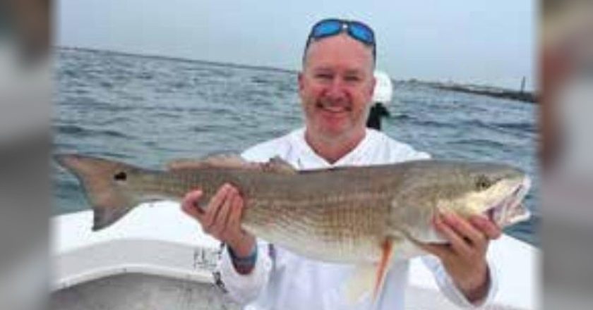 A smiling angler with sunglasses pushed up on his head, sitting on a cooler on a boat and holding a big redfish, with the inlet jetty behind him