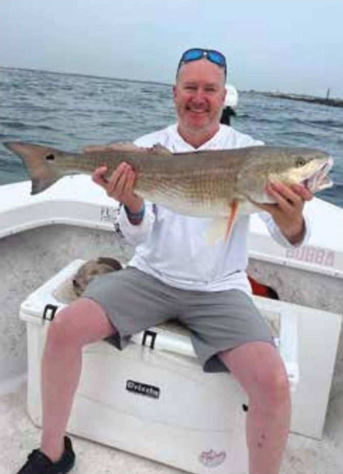 A smiling angler with sunglasses pushed up on his head, sitting on a cooler on a boat and holding a big redfish, with the inlet jetty behind him