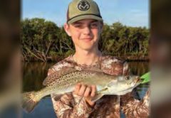 A young angler in a cap and camouflage shirt holding a spotted seatrout in front of the mangroves