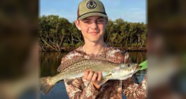 A young angler in a cap and camouflage shirt holding a spotted seatrout in front of the mangroves