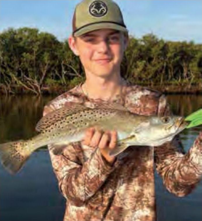 A young angler in a cap and camouflage shirt holding a spotted seatrout in front of the mangroves