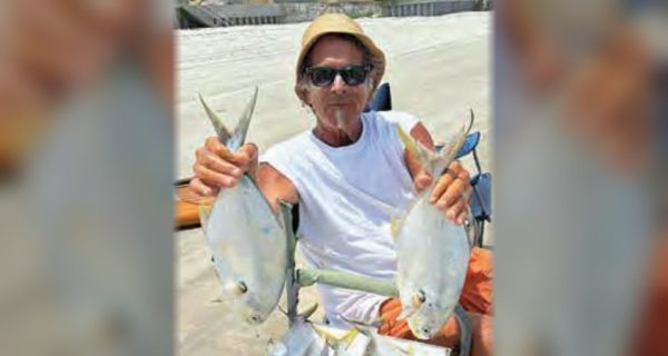 Marco Pompano holding up a pompano in each hand on the beach, with more pompano piled on a cooler in front of him