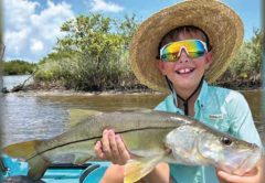 Young angler Dawson Kai McMahon in a straw hat and rainbow sunglasses holding a big snook in his kayak