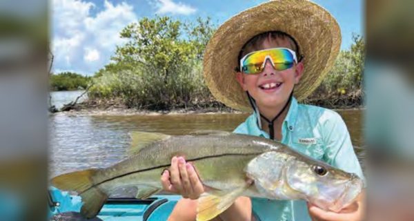 Young angler Dawson Kai McMahon in a straw hat and rainbow sunglasses holding a big snook in his kayak