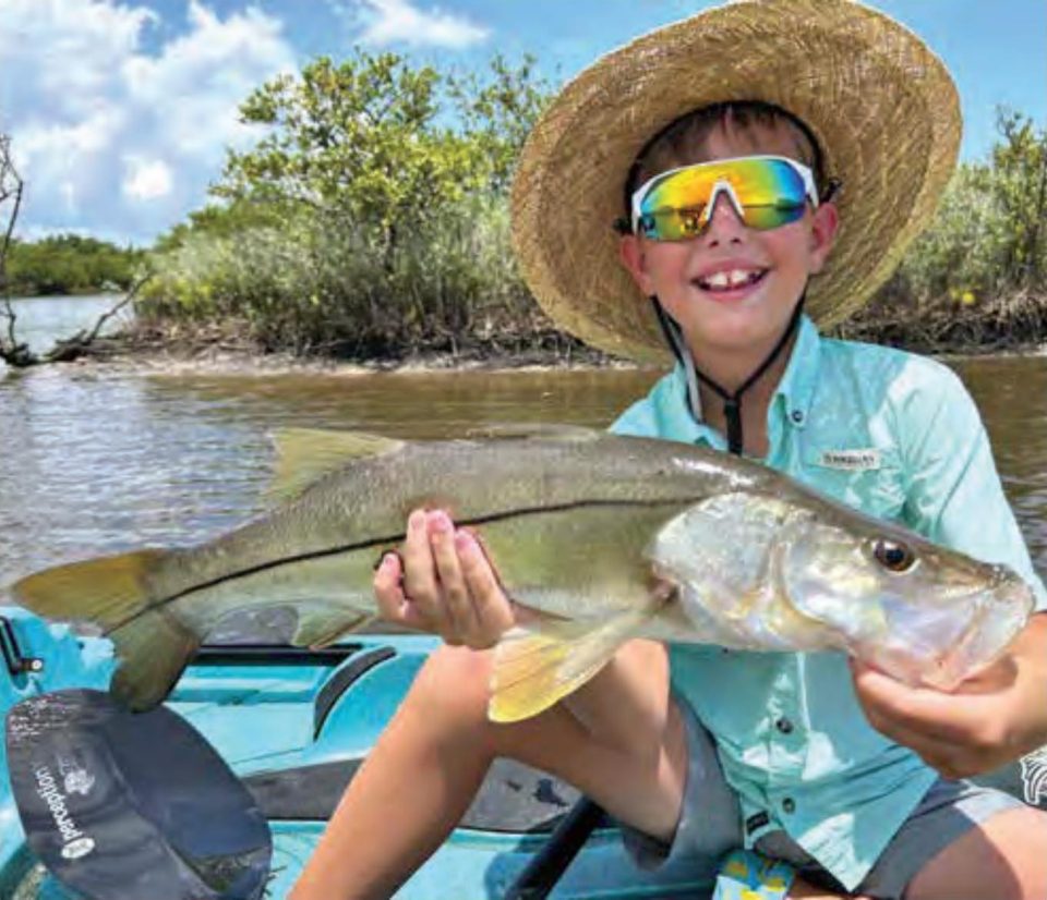 Young angler Dawson Kai McMahon in a straw hat and rainbow sunglasses holding a big snook in his kayak