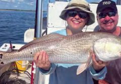 A smiling woman in a bucket hat and a man in a cap holding a big redfish on a boat