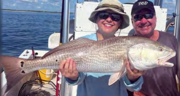 A smiling woman in a bucket hat and a man in a cap holding a big redfish on a boat