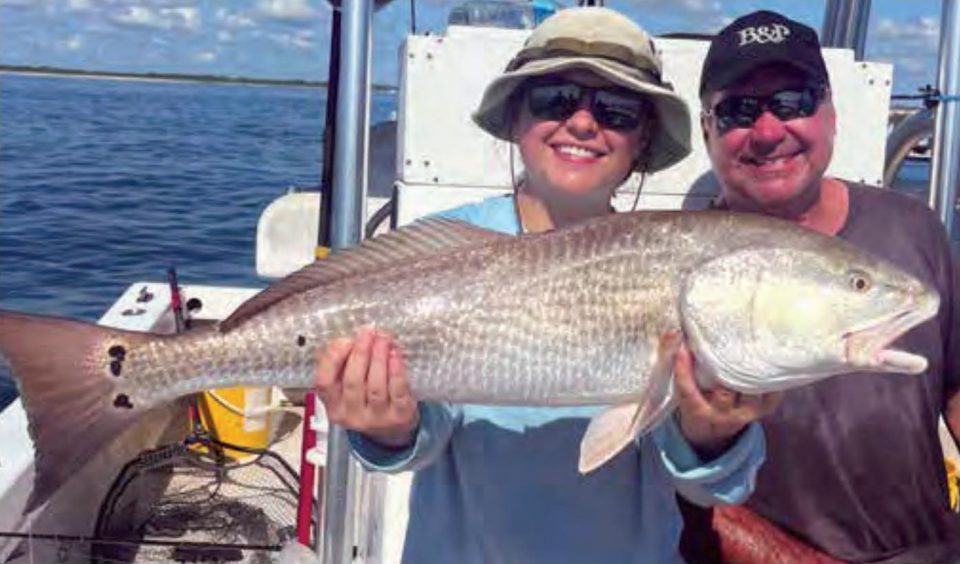 A smiling woman in a bucket hat and a man in a cap holding a big redfish on a boat