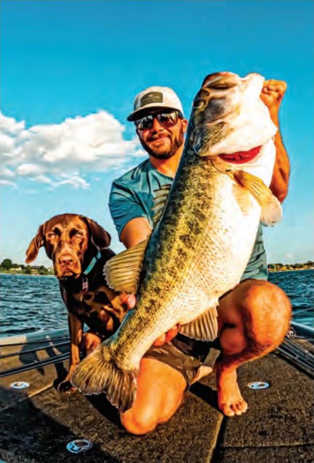Capt. Tyler Woolcott holding up a huge largemouth bass on the deck of his boat next to his chocolate lab