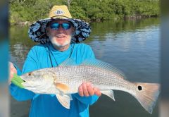 A smiling angler in a wide-brimmed sun hat and sunglasses holding a redfish in front of the mangroves