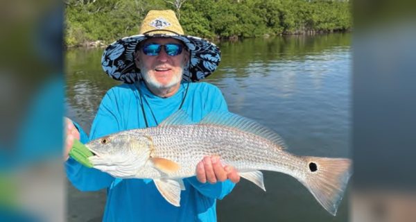 A smiling angler in a wide-brimmed sun hat and sunglasses holding a redfish in front of the mangroves