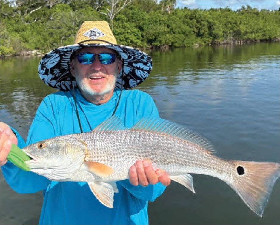 A smiling angler in a wide-brimmed sun hat and sunglasses holding a redfish in front of the mangroves